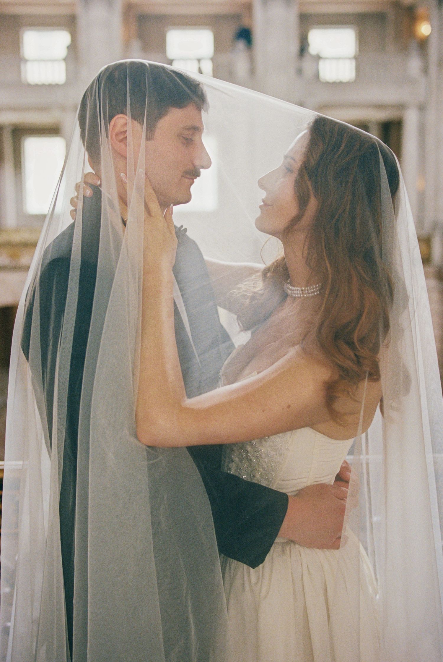 Bride and groom share an intimate moment under a flowing white wedding veil in an elegant venue.