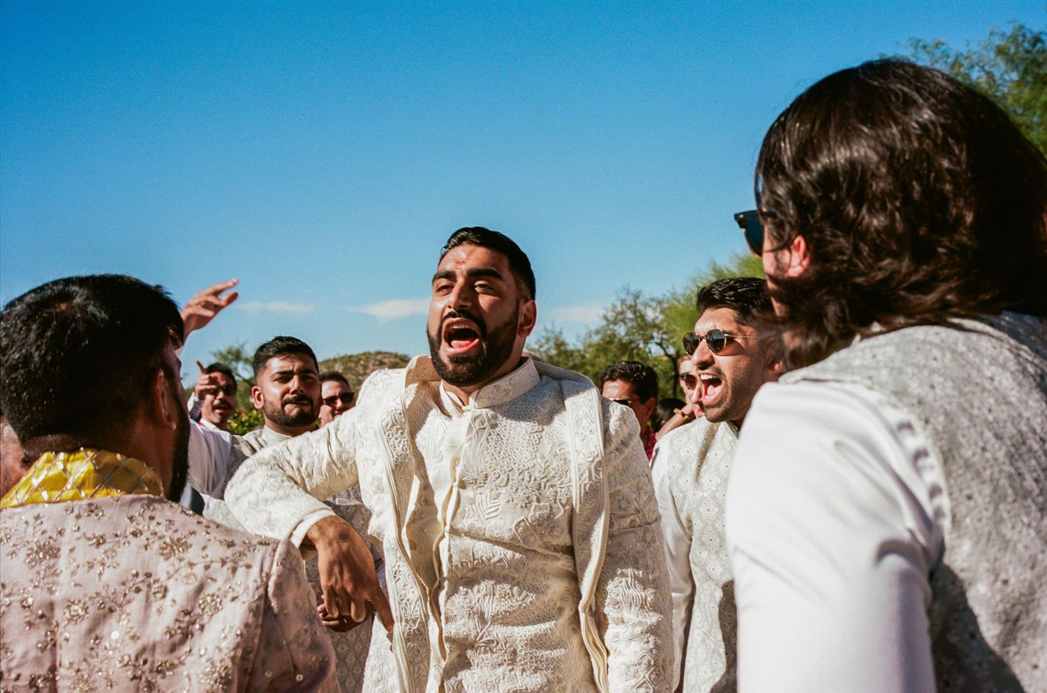 Wedding party members in white traditional attire celebrate with enthusiastic expressions and gestures.
