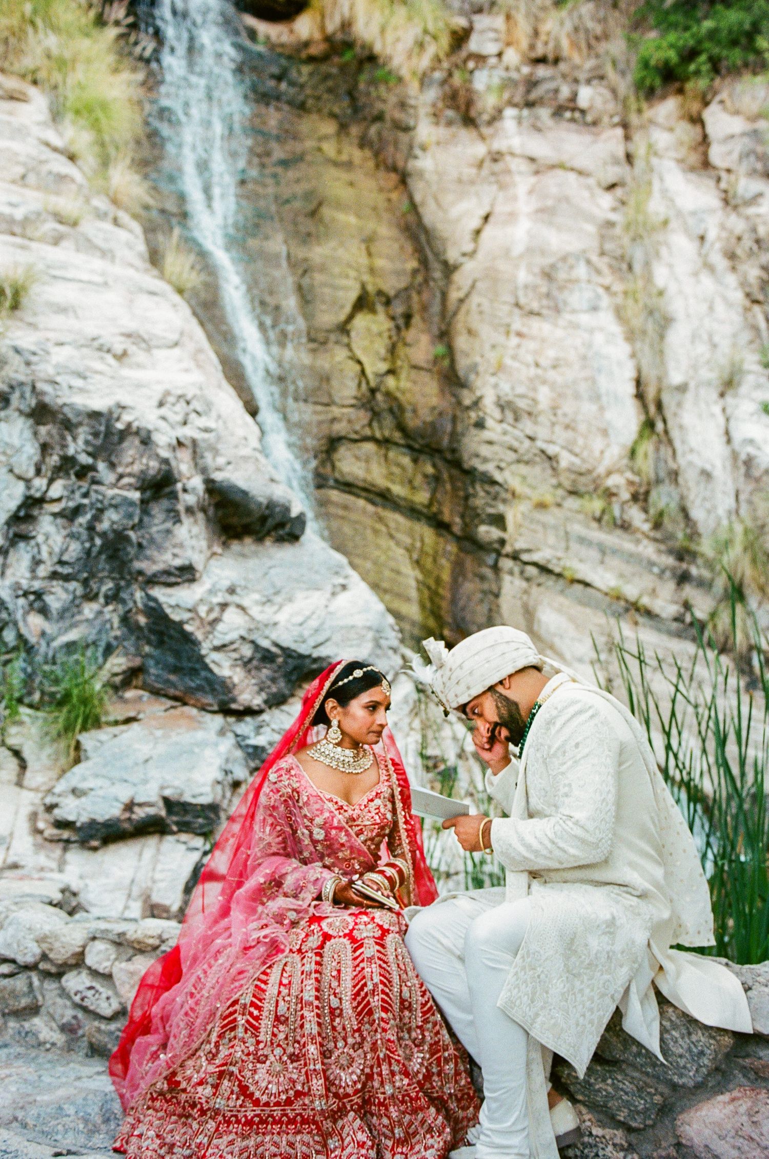 Couple in traditional Indian wedding attire sitting by scenic waterfall.