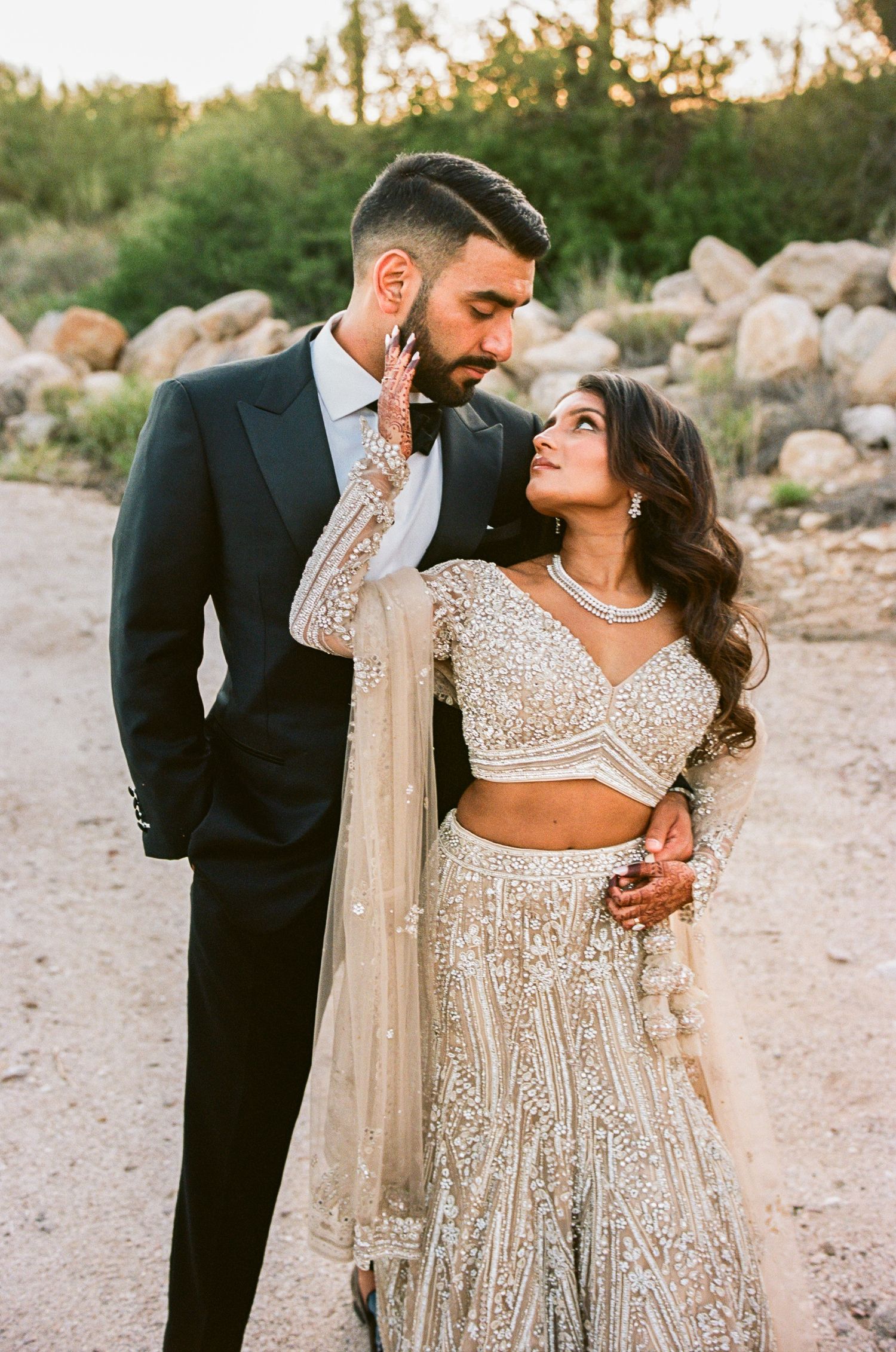 A glamorous couple in formal attire sharing a romantic moment in a desert setting at sunset.