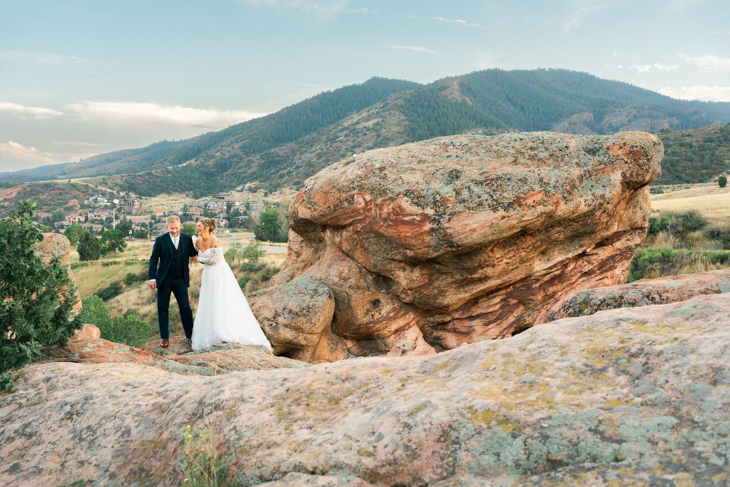 Bride and groom holding hands on rocky mountain overlook with scenic Colorado landscape and rolling hills in background.
