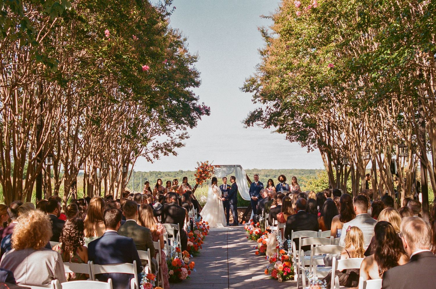 Wedding ceremony in progress under canopy of autumn trees with guests seated on white chairs.