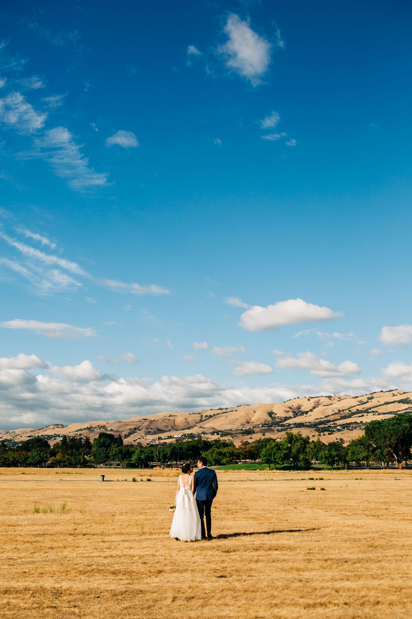 A couple stands together in a golden field beneath blue skies and rolling hills on their wedding day.