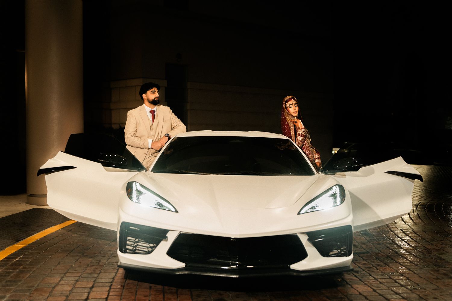 Couple in traditional attire posing with a white Corvette sports car in a dramatic nighttime setting.