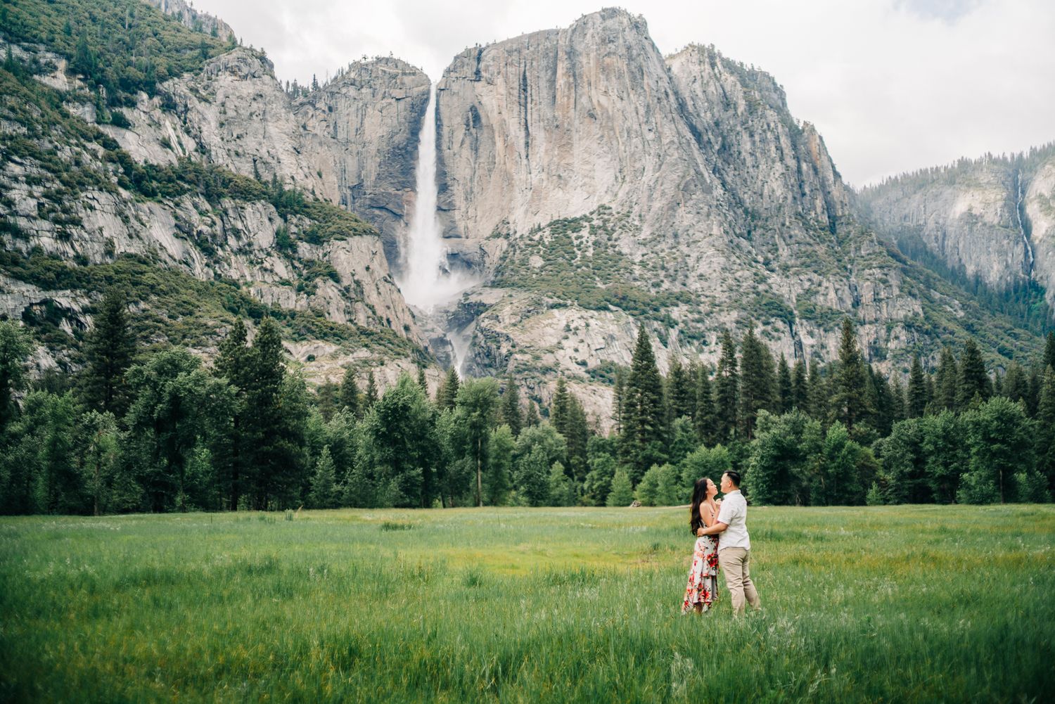 Couple embracing in Yosemite Valley meadow with Yosemite Falls and granite cliffs in background.