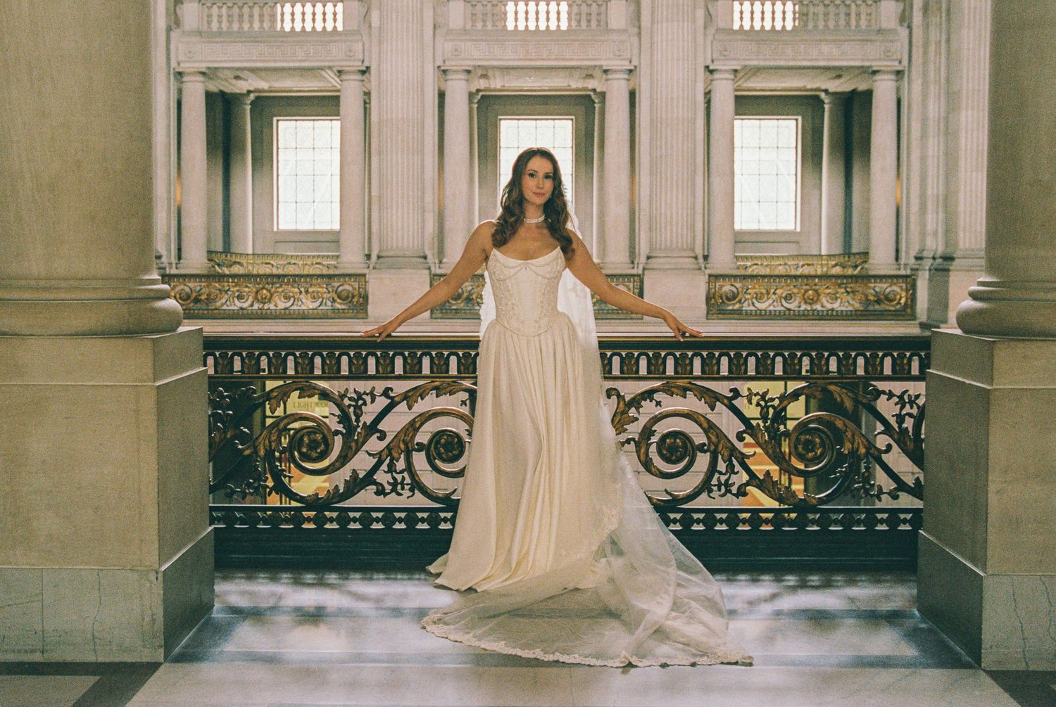 Elegant bride in a flowing white gown poses on an ornate balcony inside a grand classical building.