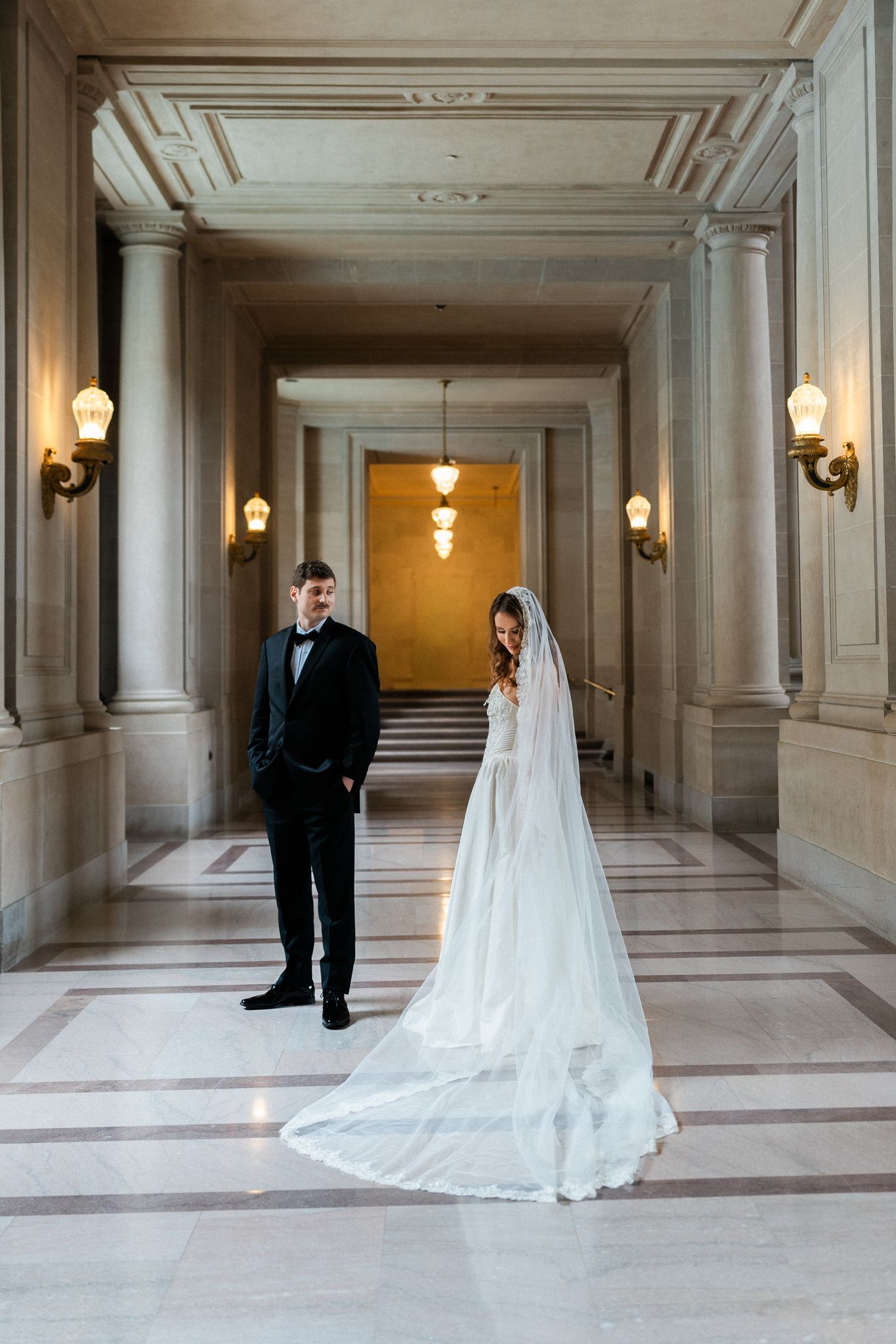 Bride in white gown with long veil and groom in black tuxedo pose in elegant marble hall with ornate columns.
