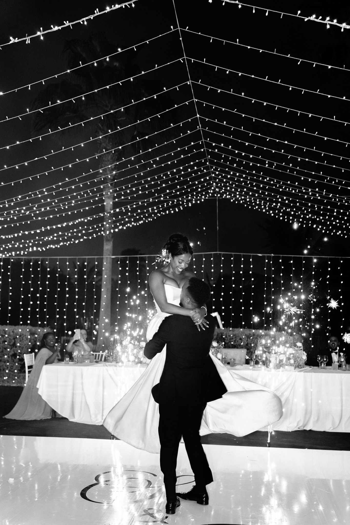Bride and groom share first dance under twinkling fairy lights at elegant wedding reception in black and white photo.