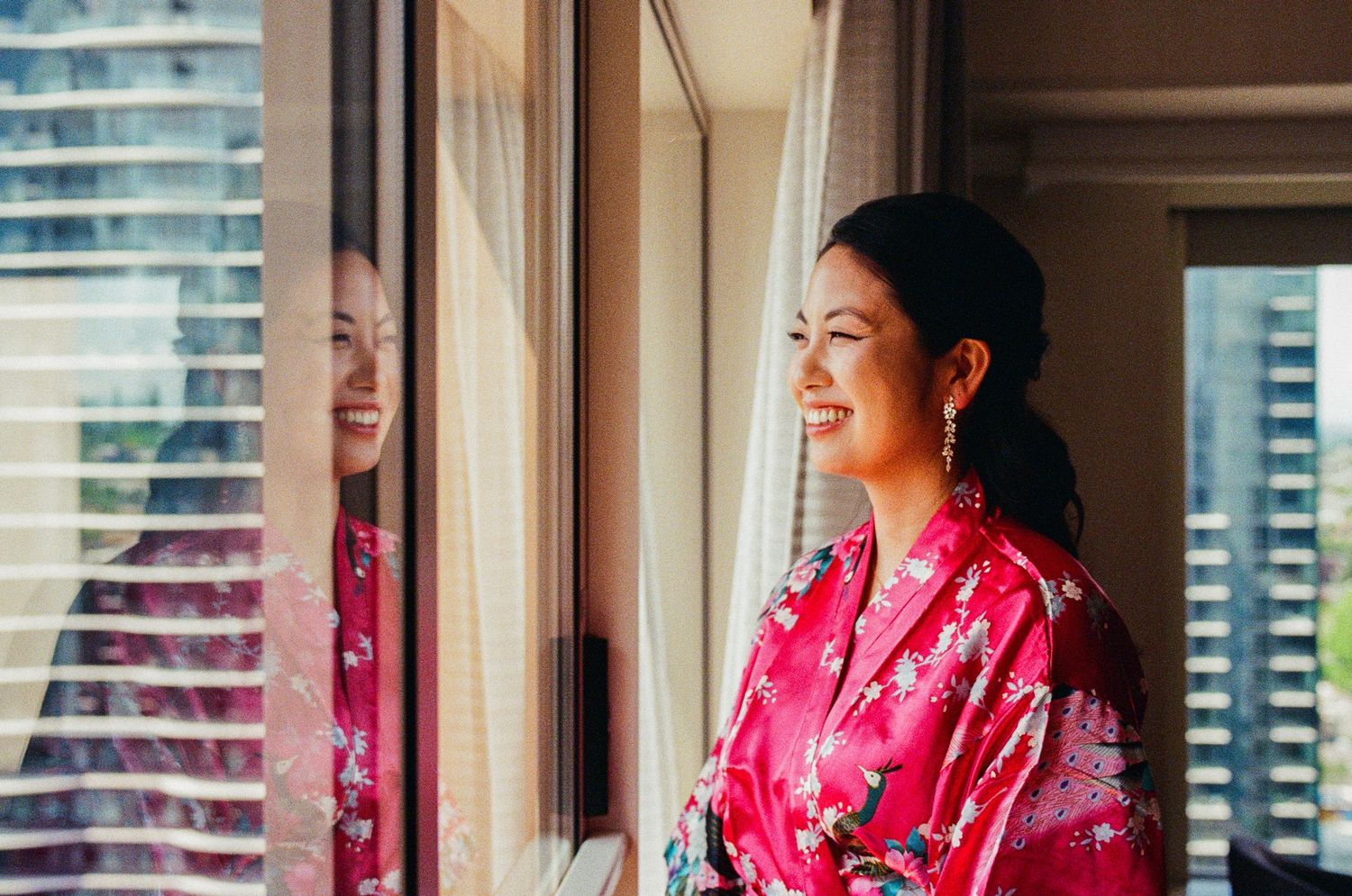 Person in pink floral robe smiling while looking out window in city apartment.