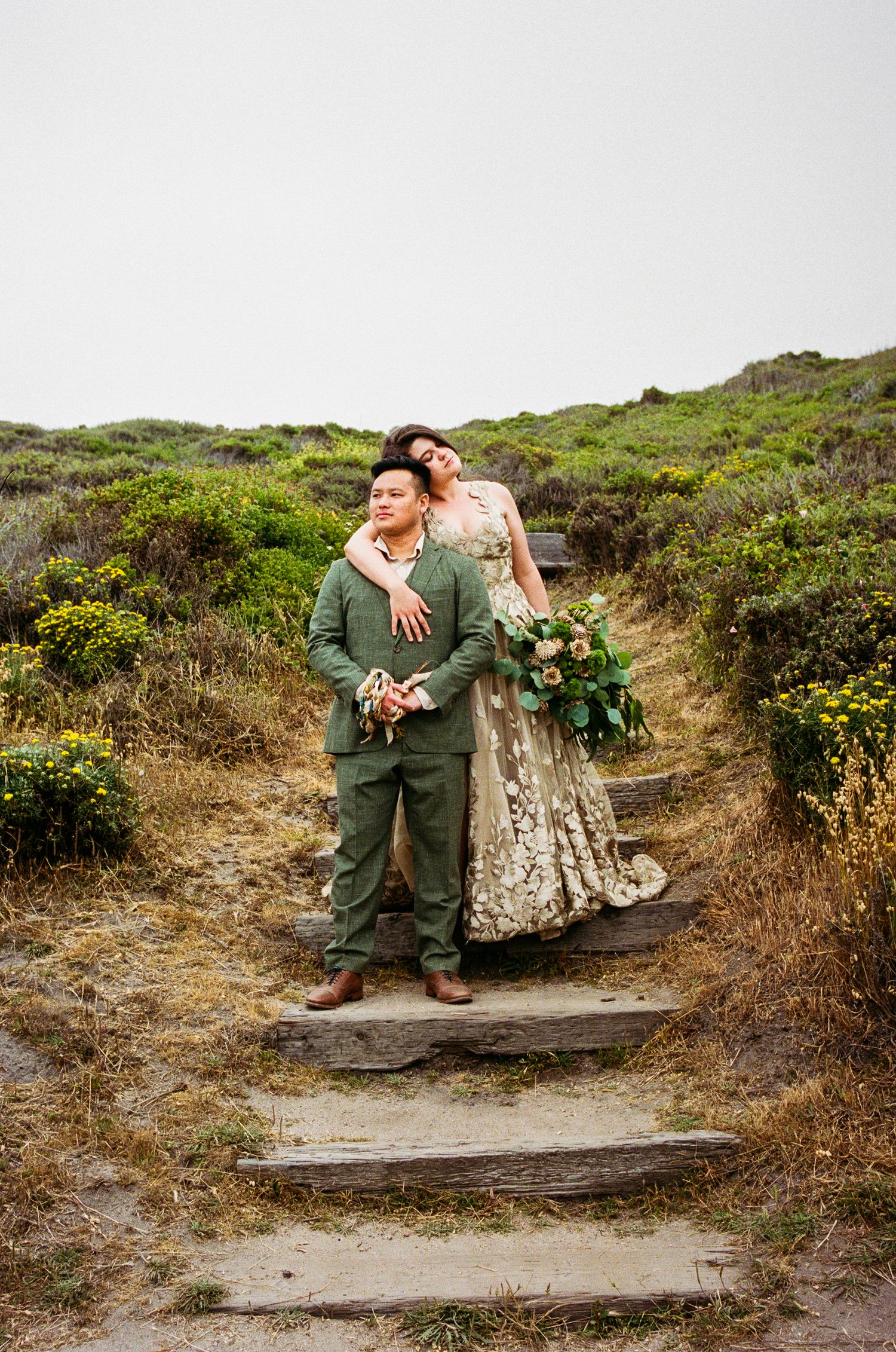 A wedding couple poses on stone steps surrounded by coastal vegetation.