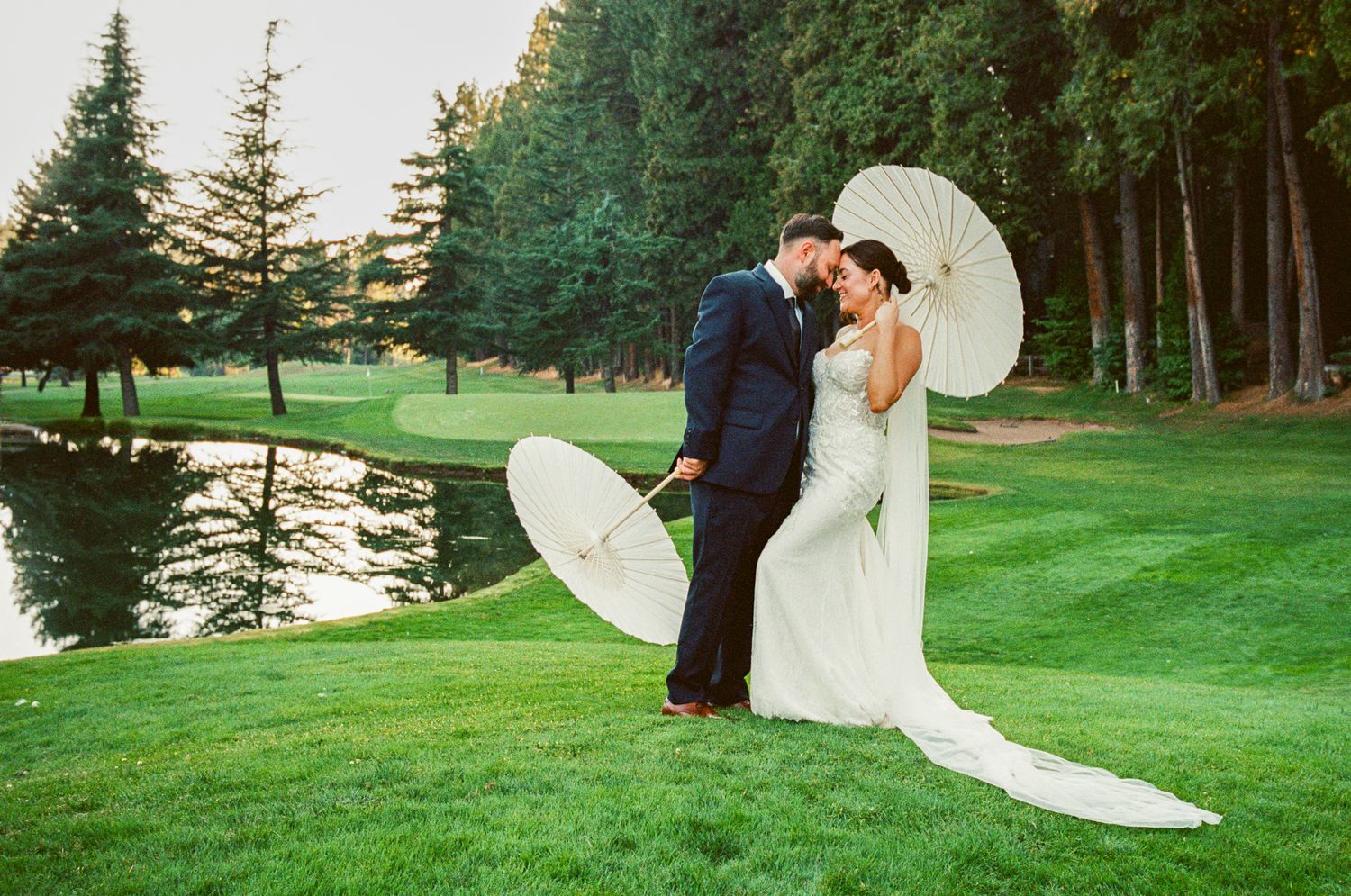 A couple shares a tender moment during their wedding photoshoot on a golf course with white parasol.