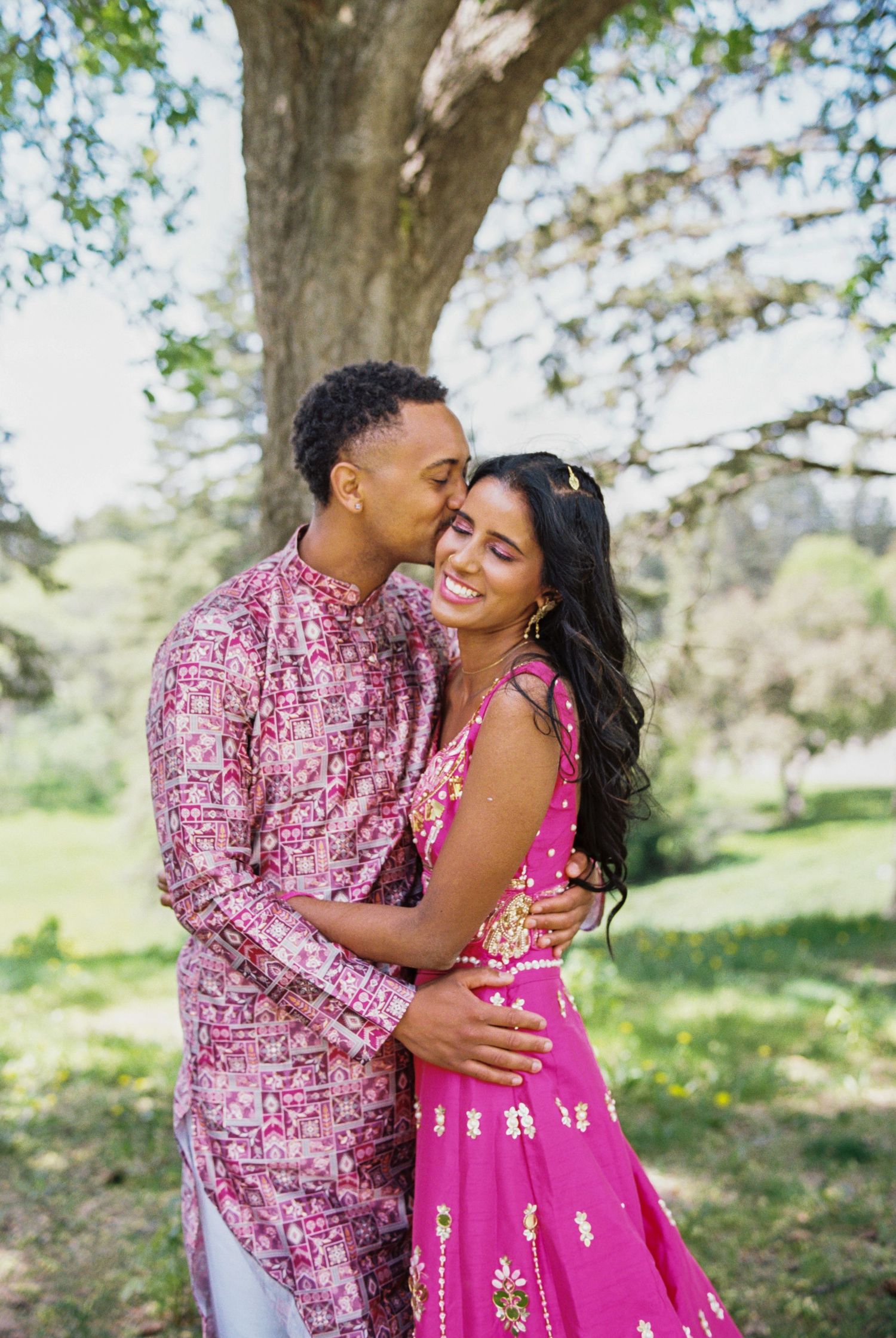 Couple in matching pink outfits sharing a romantic moment under a large tree in a lush green garden.