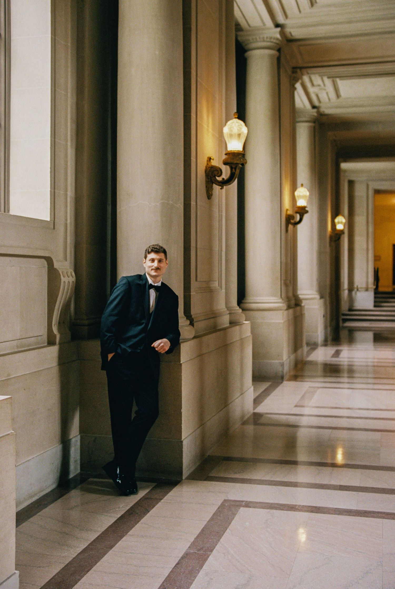 Young man in tuxedo leaning against ornate marble columns in elegant hallway with wall sconces.