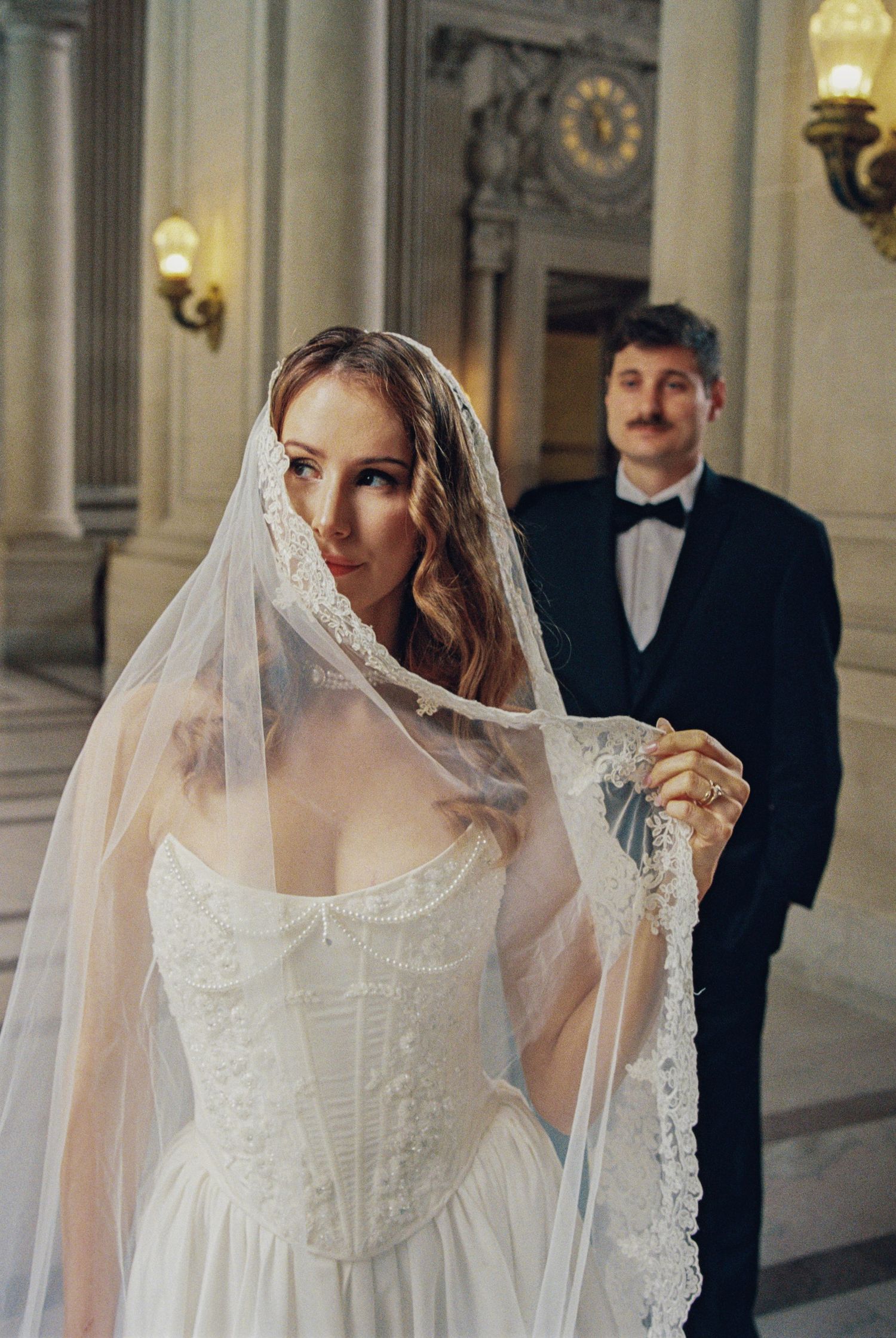 Bride in lace corset wedding dress lifts veil with groom in tuxedo standing behind in elegant venue.