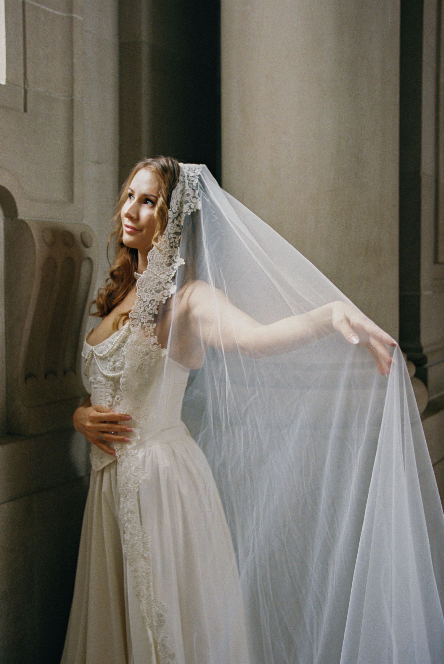 Bride in lace wedding dress holding flowing tulle veil beside stone columns in elegant church setting.