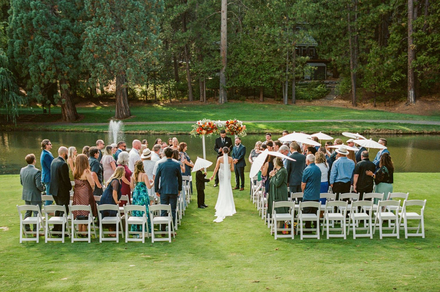 An outdoor wedding ceremony with guests seated on white chairs by a serene lake.