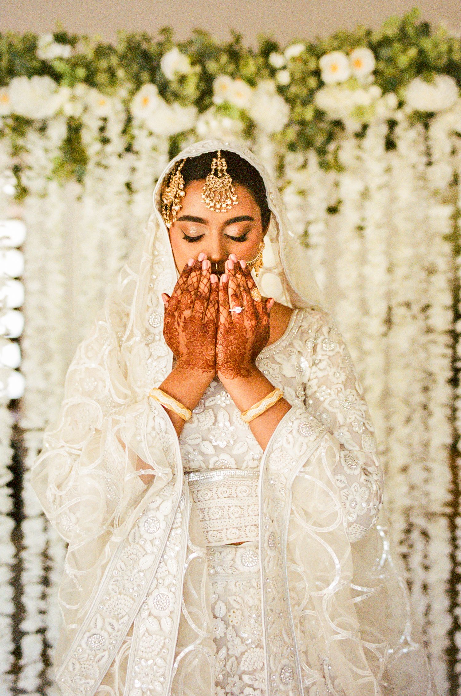 Traditional bridal portrait with white floral backdrop and henna-decorated hands.
