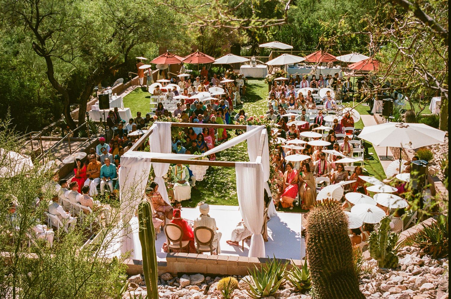 Outdoor wedding reception setup with white tables and umbrellas in a natural setting.