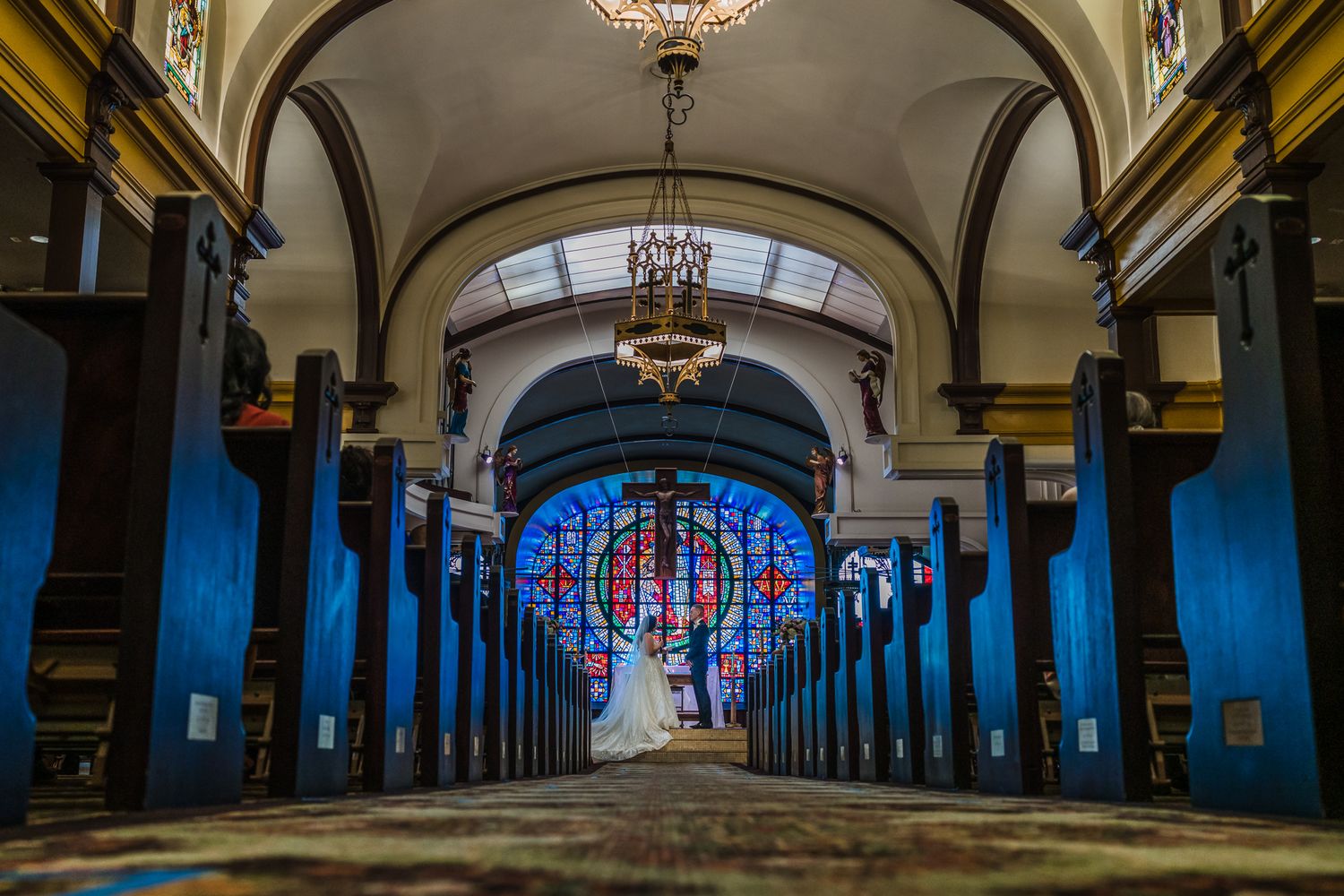 Elegant church interior with blue pews, arched ceilings, chandelier, and stunning stained glass window at the altar.