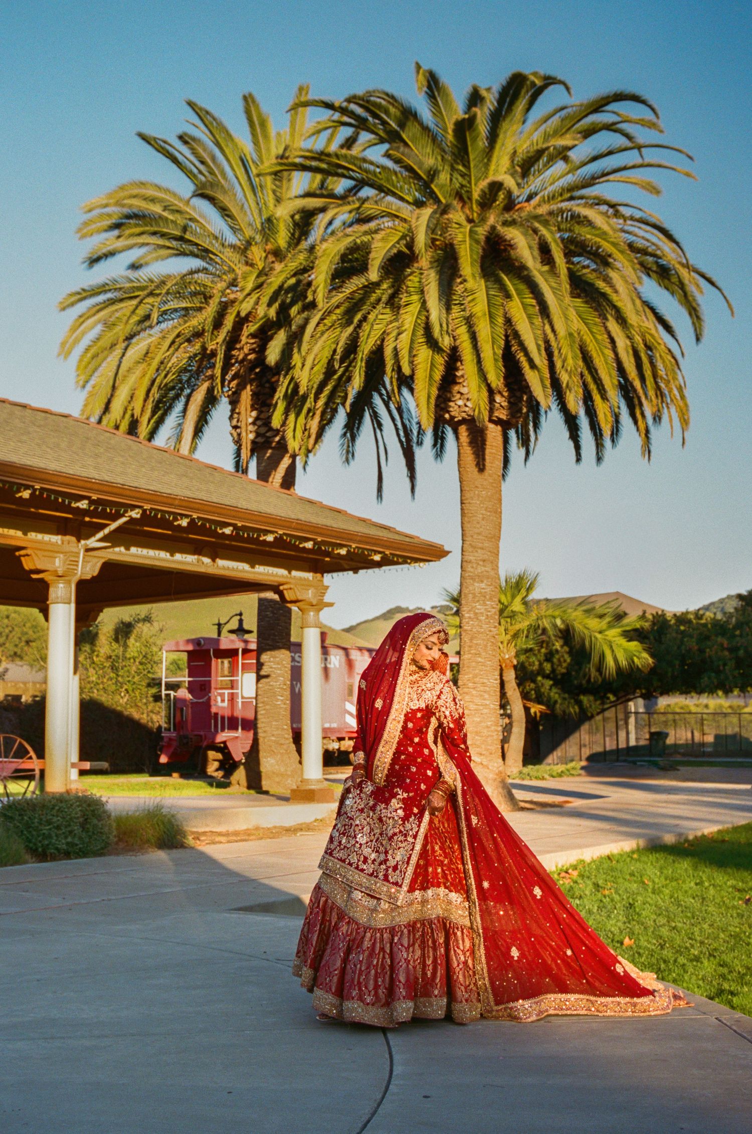 Ornate red and gold lehenga dress photographed against palm trees and outdoor architecture.