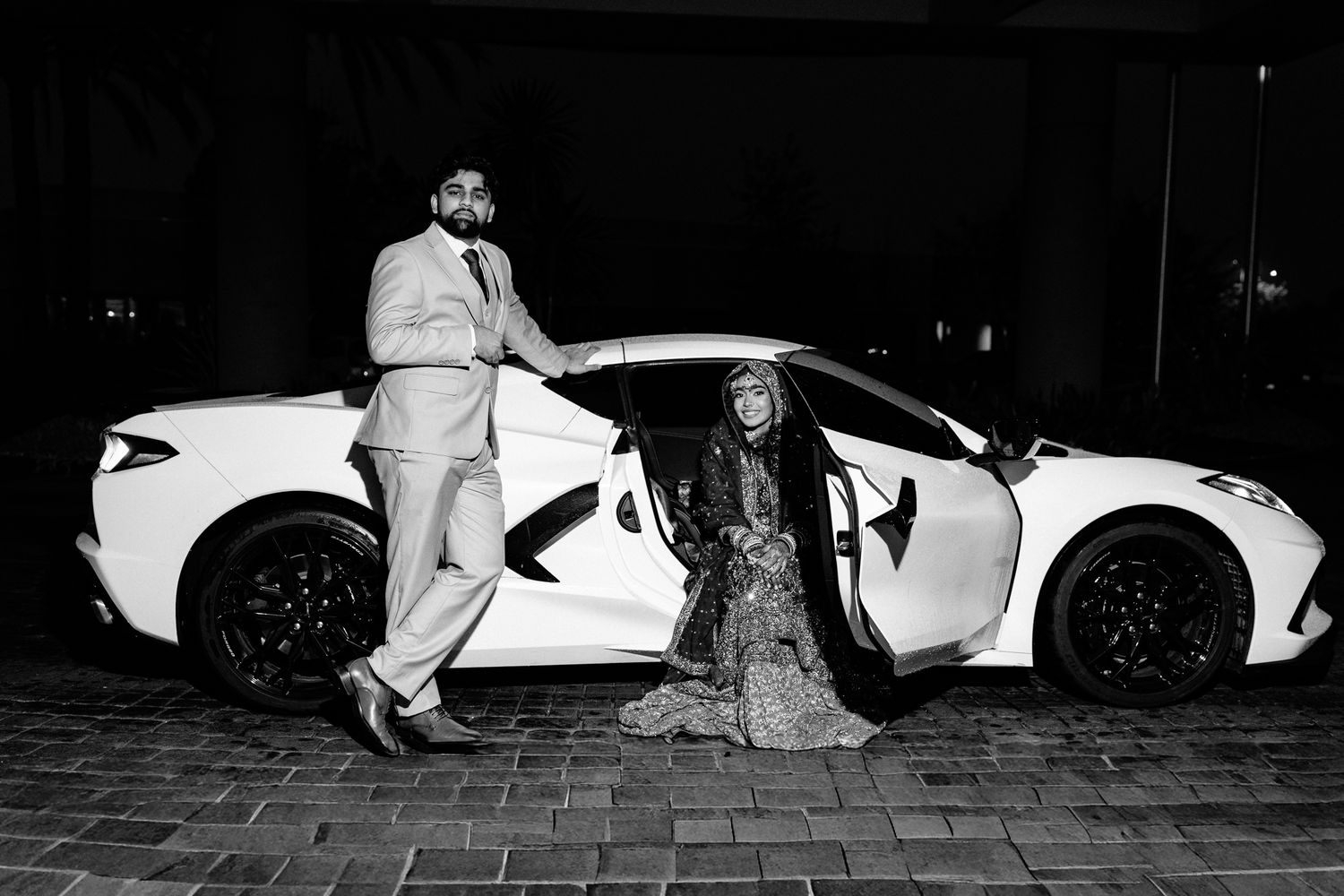 Couple in formal attire posing with a white sports car in a black and white photo.