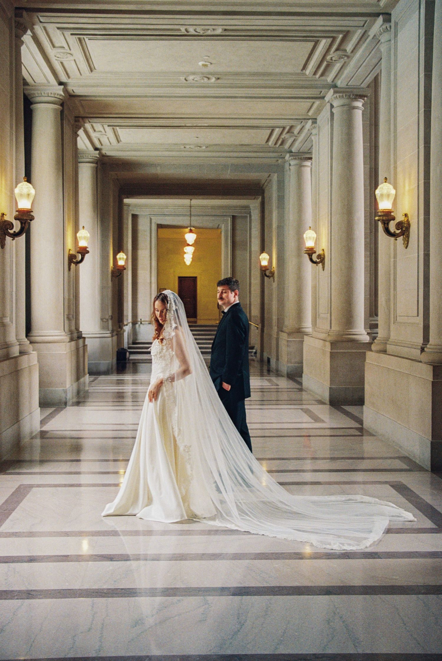 Bride and groom stand in elegant marble hallway with tall columns and warm wall sconces at San Francisco City Hall wedding.