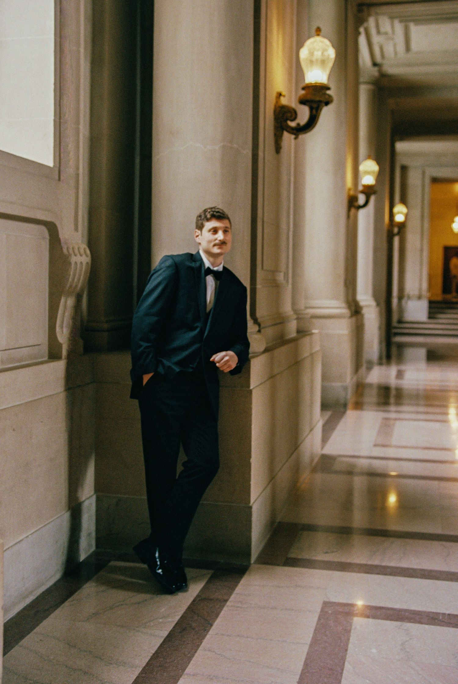 Young man in black tuxedo leaning against ornate column in elegant hallway with wall sconces.