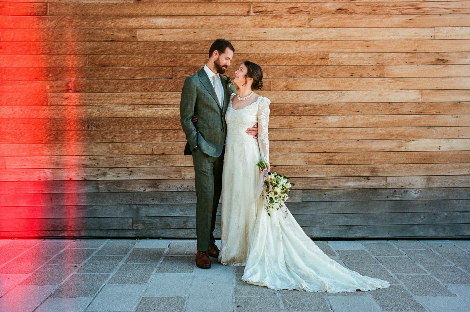 A romantic wedding couple poses together against a rustic wooden wall with dramatic red lighting.