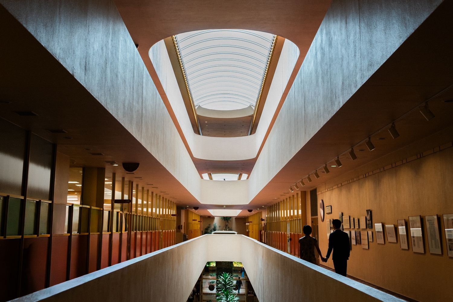 Modern building interior hallway with skylight ceiling, wood-paneled walls, and two silhouetted figures walking.