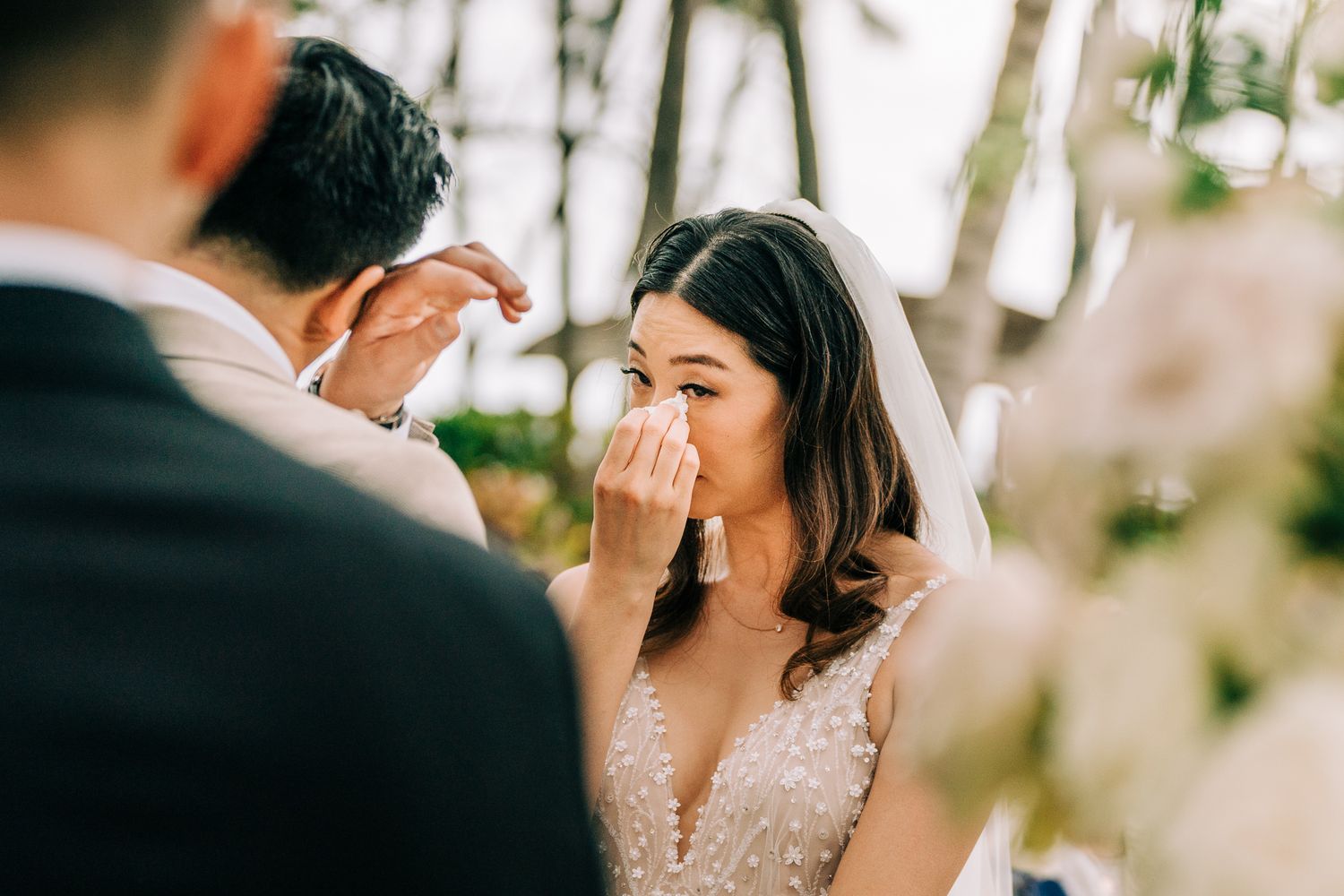 Emotional bride crying during outdoor wedding ceremony, wiping tears with tissue while groom stands before her.