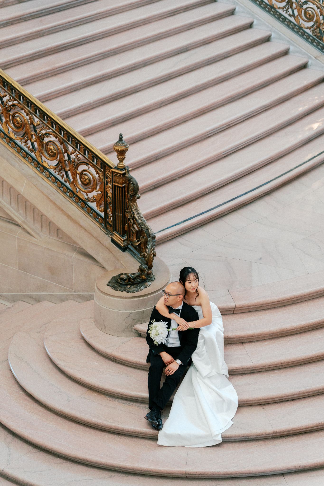 Bride and groom sitting on grand marble staircase with ornate gold railing, bride holding white bouquet.