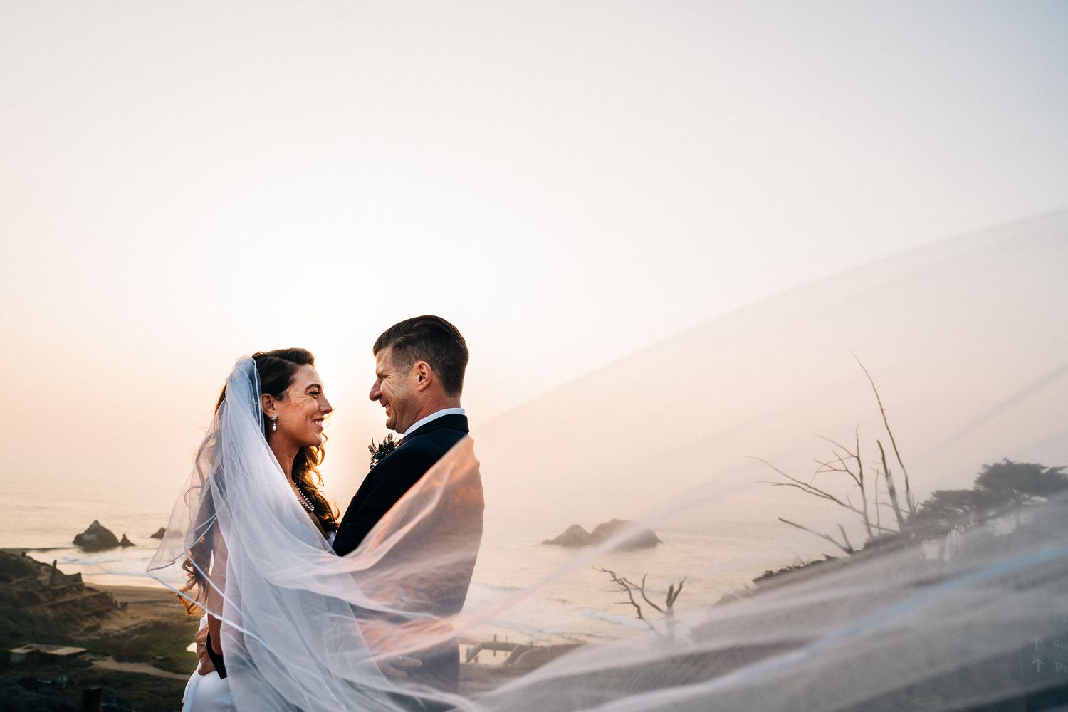 A romantic wedding couple embraces on a sandy beach during golden hour with flowing white veil in the breeze.