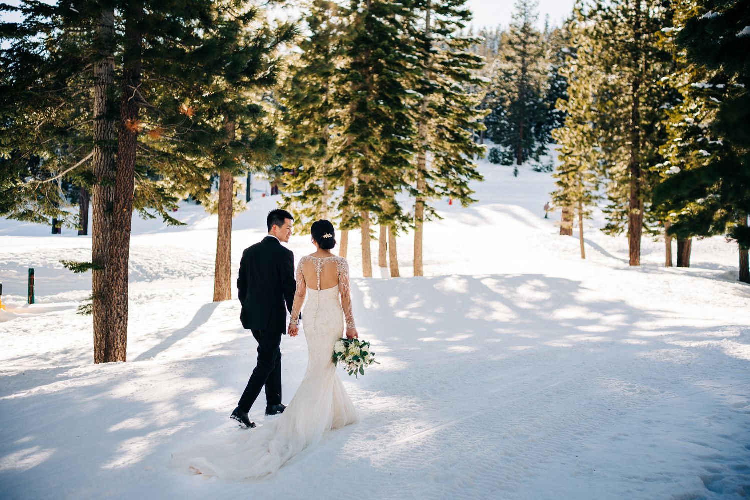 A couple walks hand in hand through a snowy forest path surrounded by tall evergreen trees in winter.