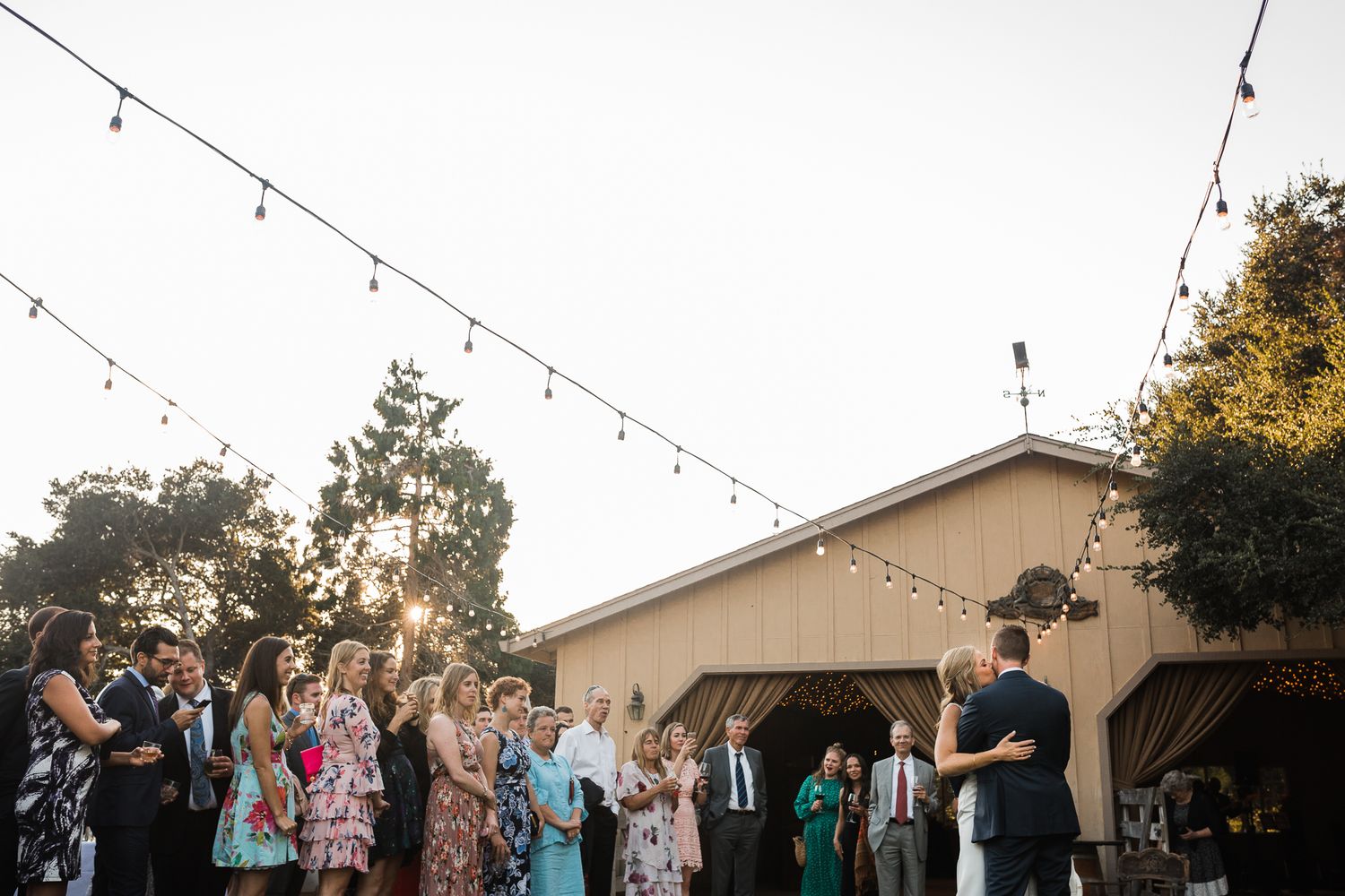 Outdoor wedding reception venue with string lights and guests mingling at dusk.