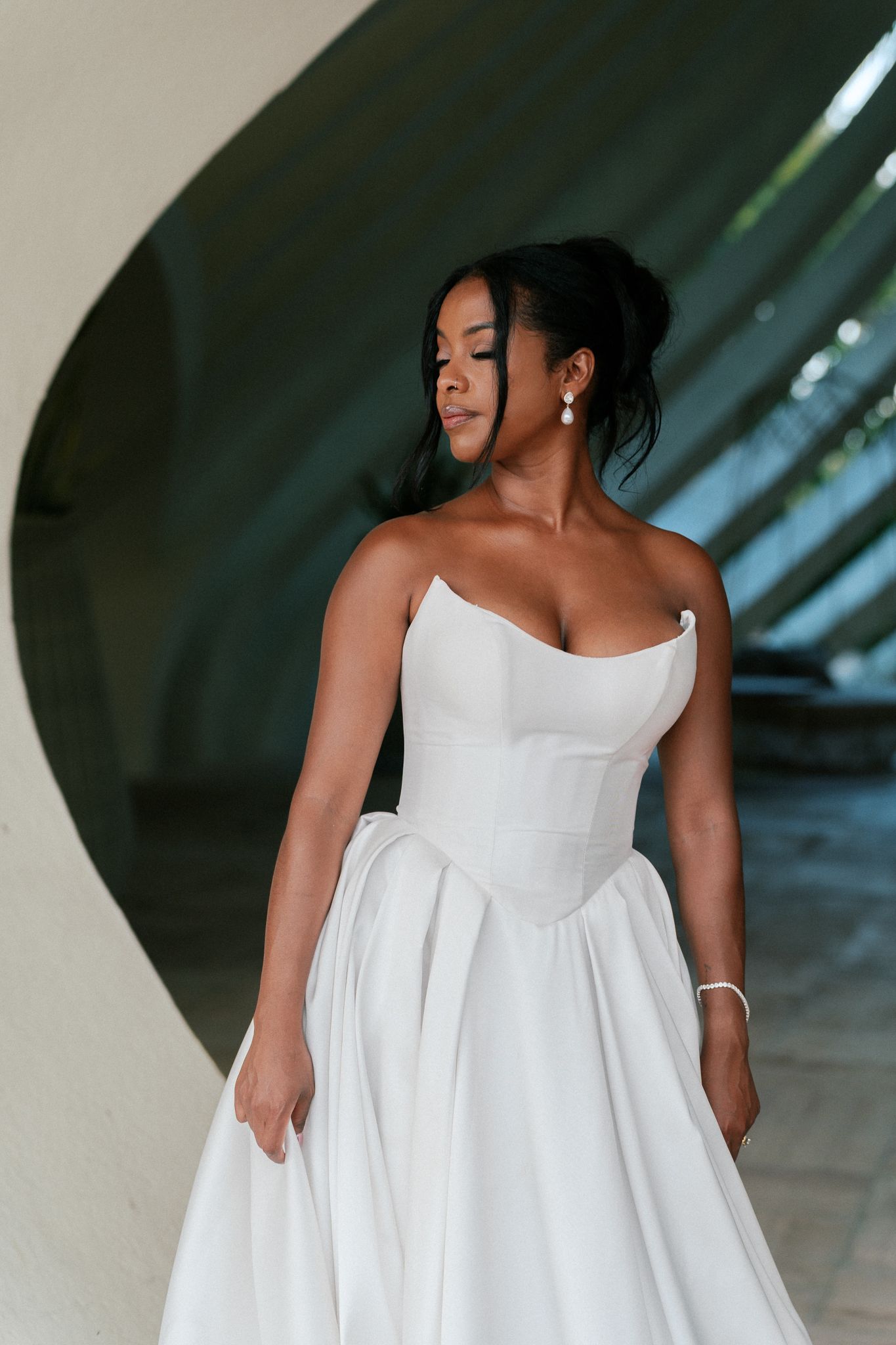 Elegant bride in strapless white ball gown with pearl earrings poses gracefully in modern architectural setting.