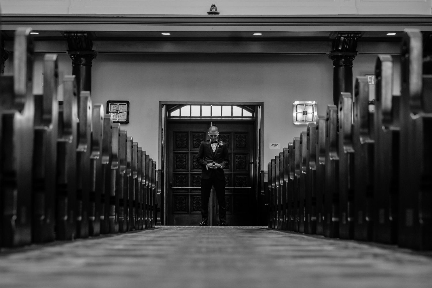 Black and white photo of a person standing alone at the end of church pews in a formal suit.