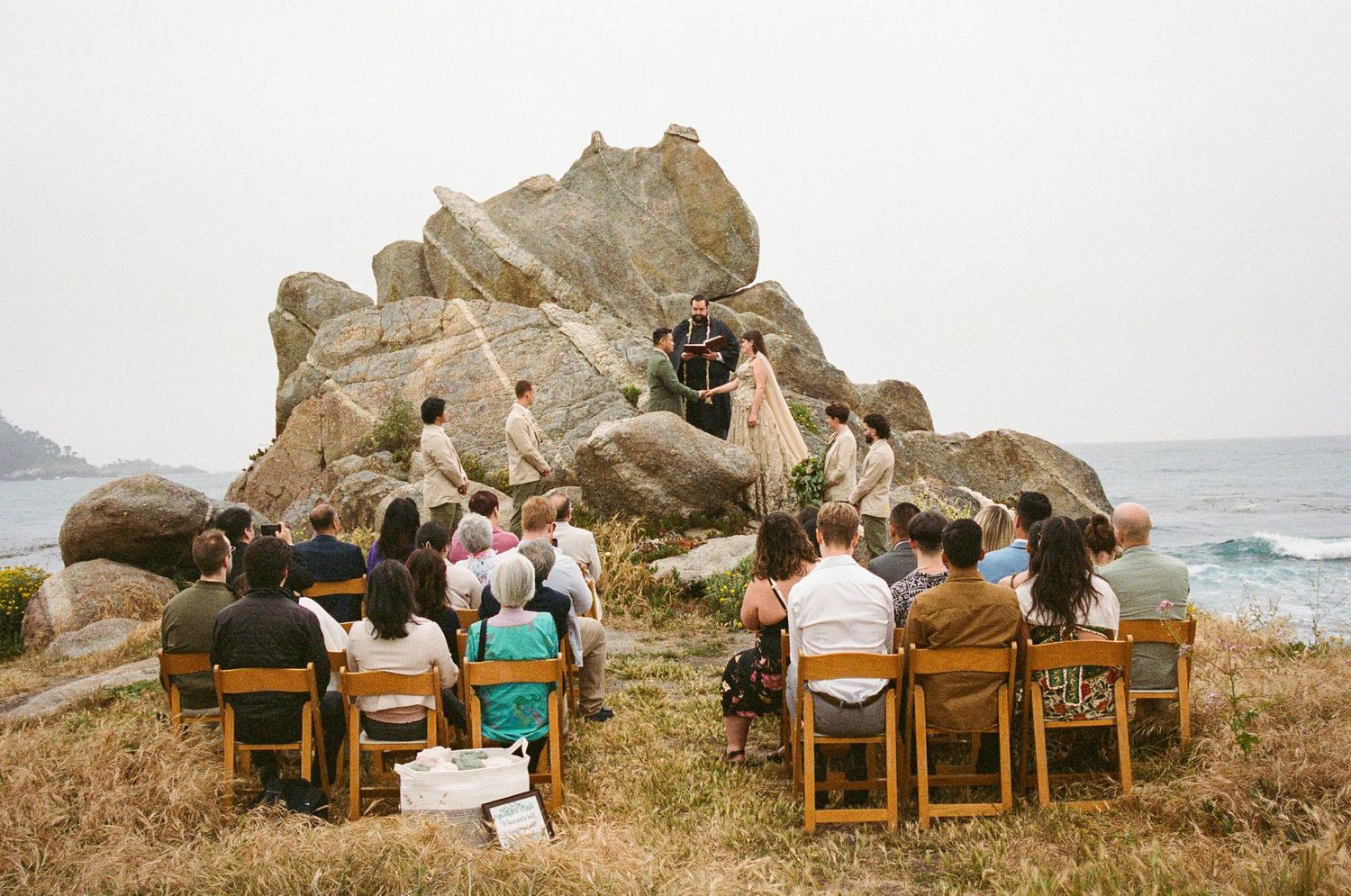Wedding party sits on coastal rocks overlooking the ocean during sunset.