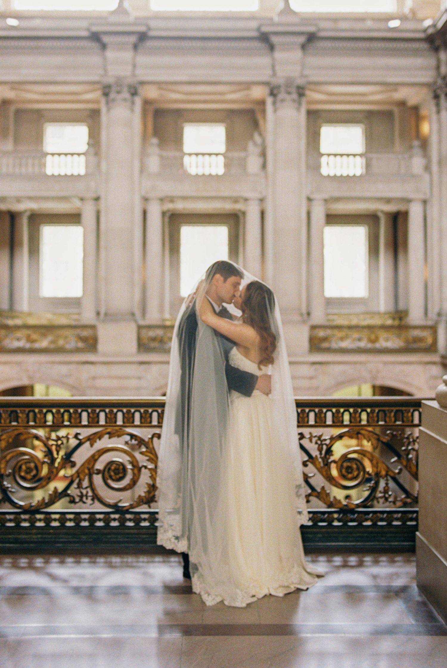 Bride and groom embrace in a grand hall with ornate gold railings, bride wearing a flowing white gown and veil.