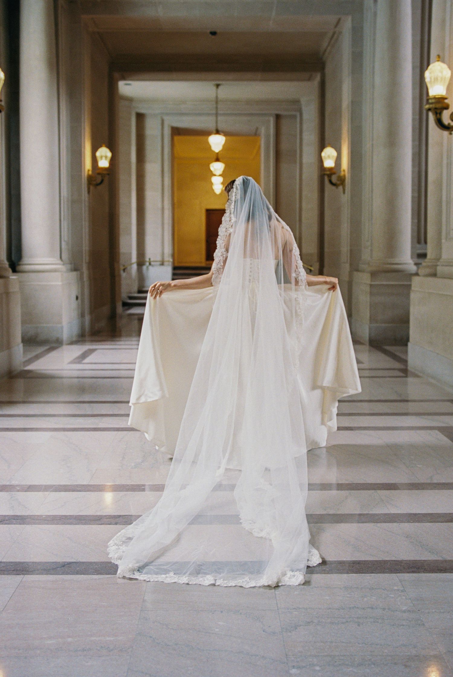 Bride in a white wedding gown and long lace-trimmed veil stands in an elegant marble hallway.