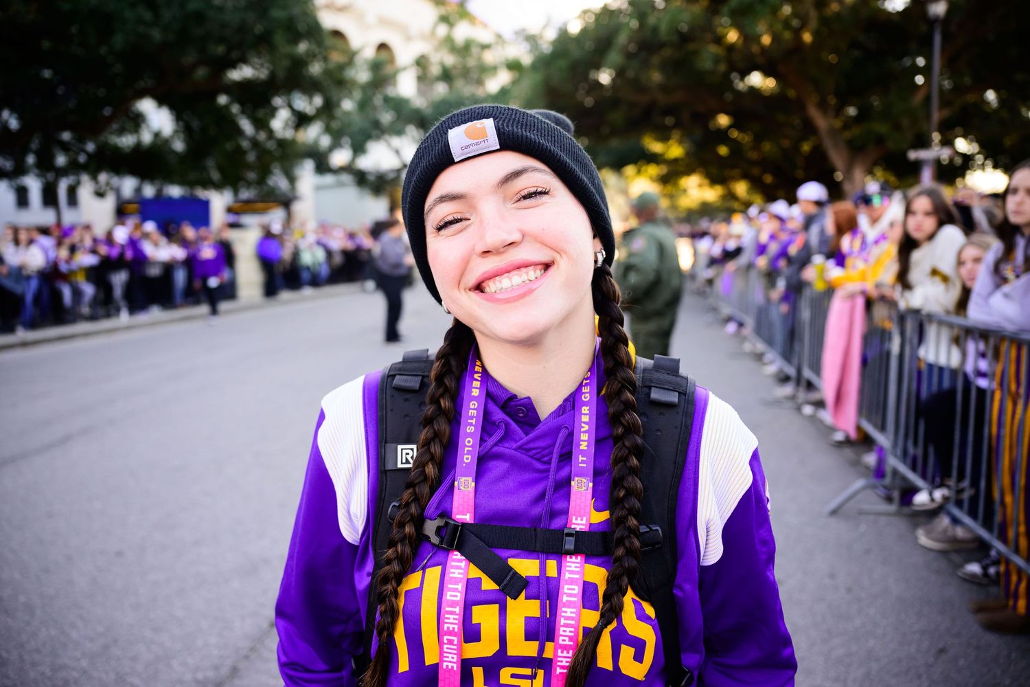 Fan wearing purple LSU hoodie and beanie smiling at outdoor sporting event.