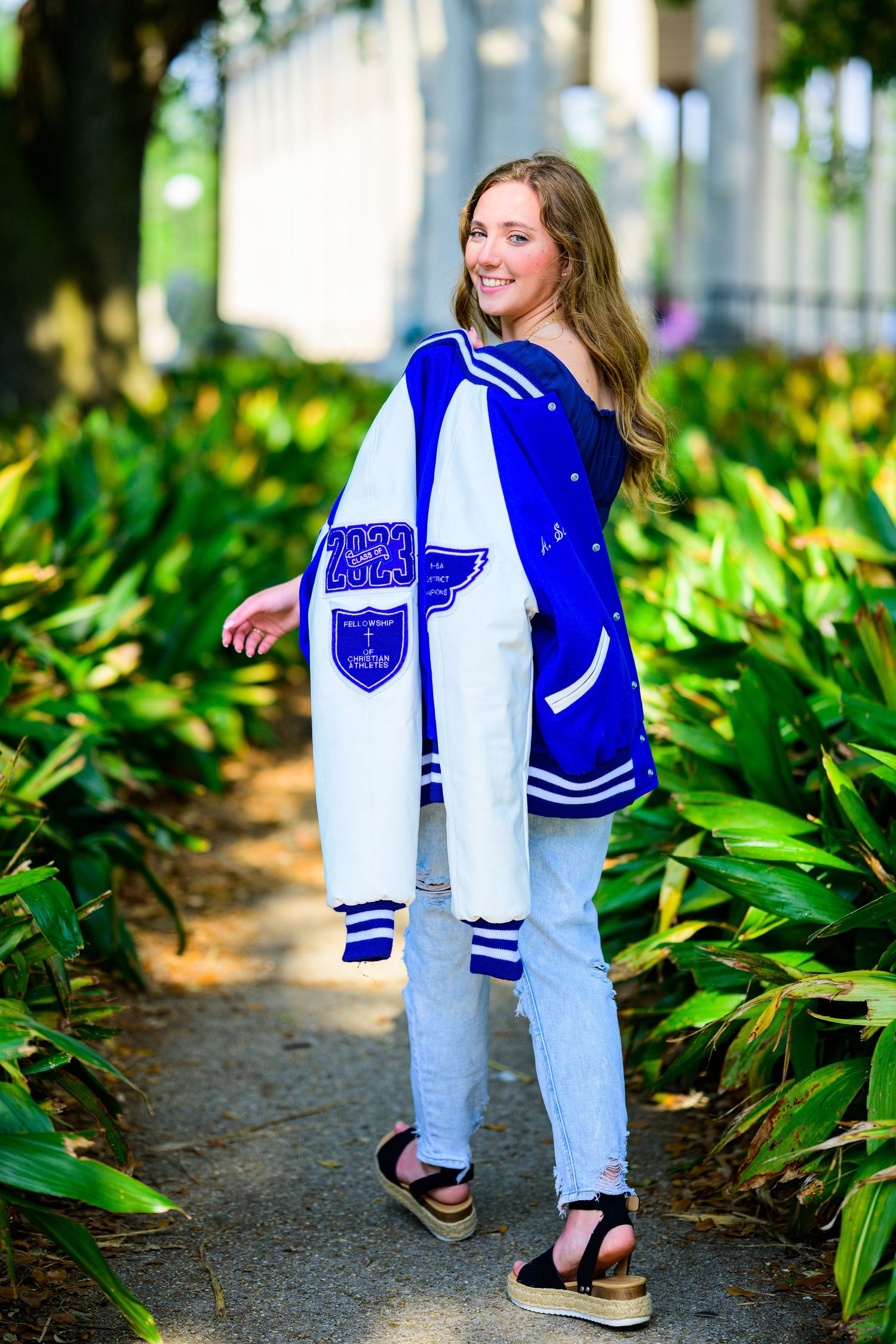 Student wearing blue and white varsity jacket poses on garden path surrounded by tropical plants.