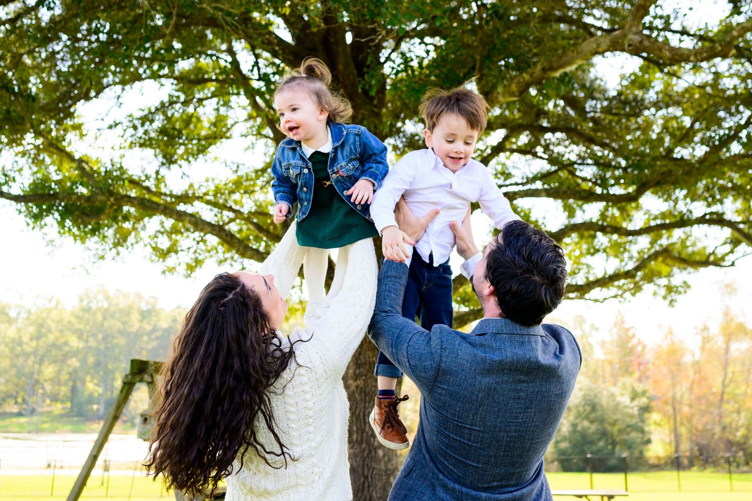 Parents playfully lifting children in the air under large tree in outdoor setting.