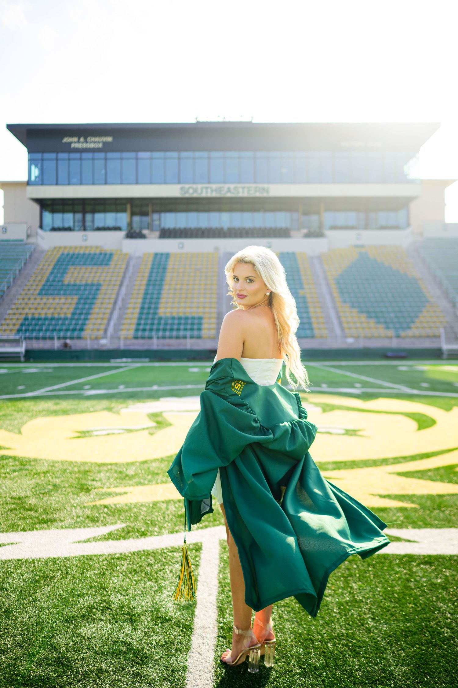 Blonde woman in green graduation gown poses on football field at Southeastern Louisiana University stadium.