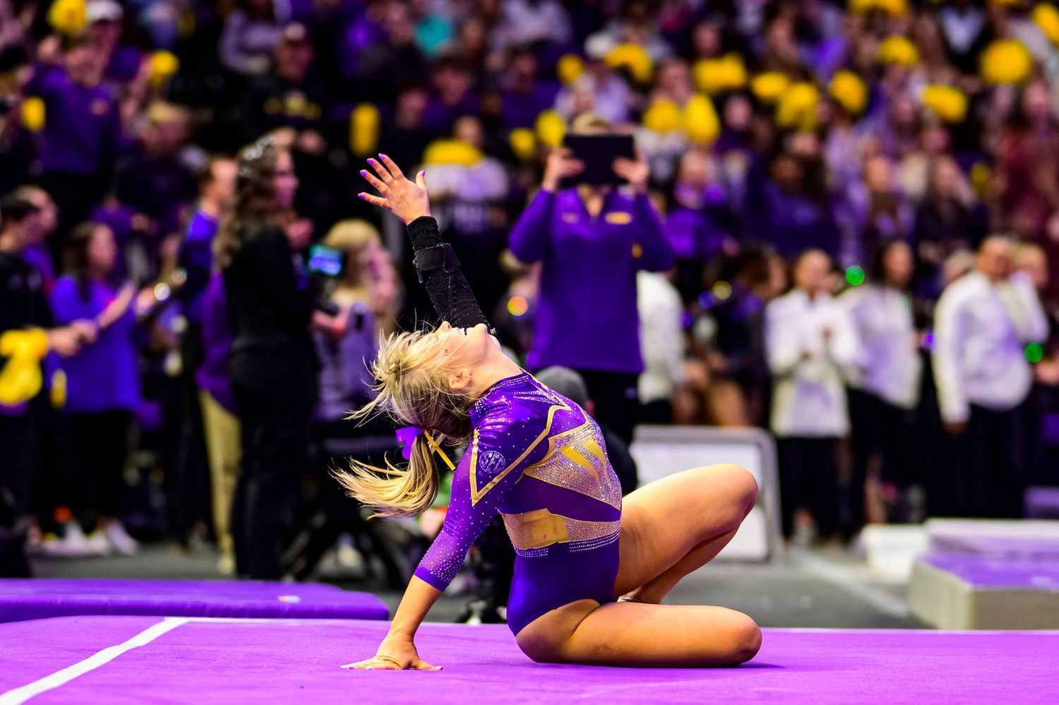 A gymnast in purple leotard performs a dynamic floor routine at a collegiate competition with purple-clad fans in background.