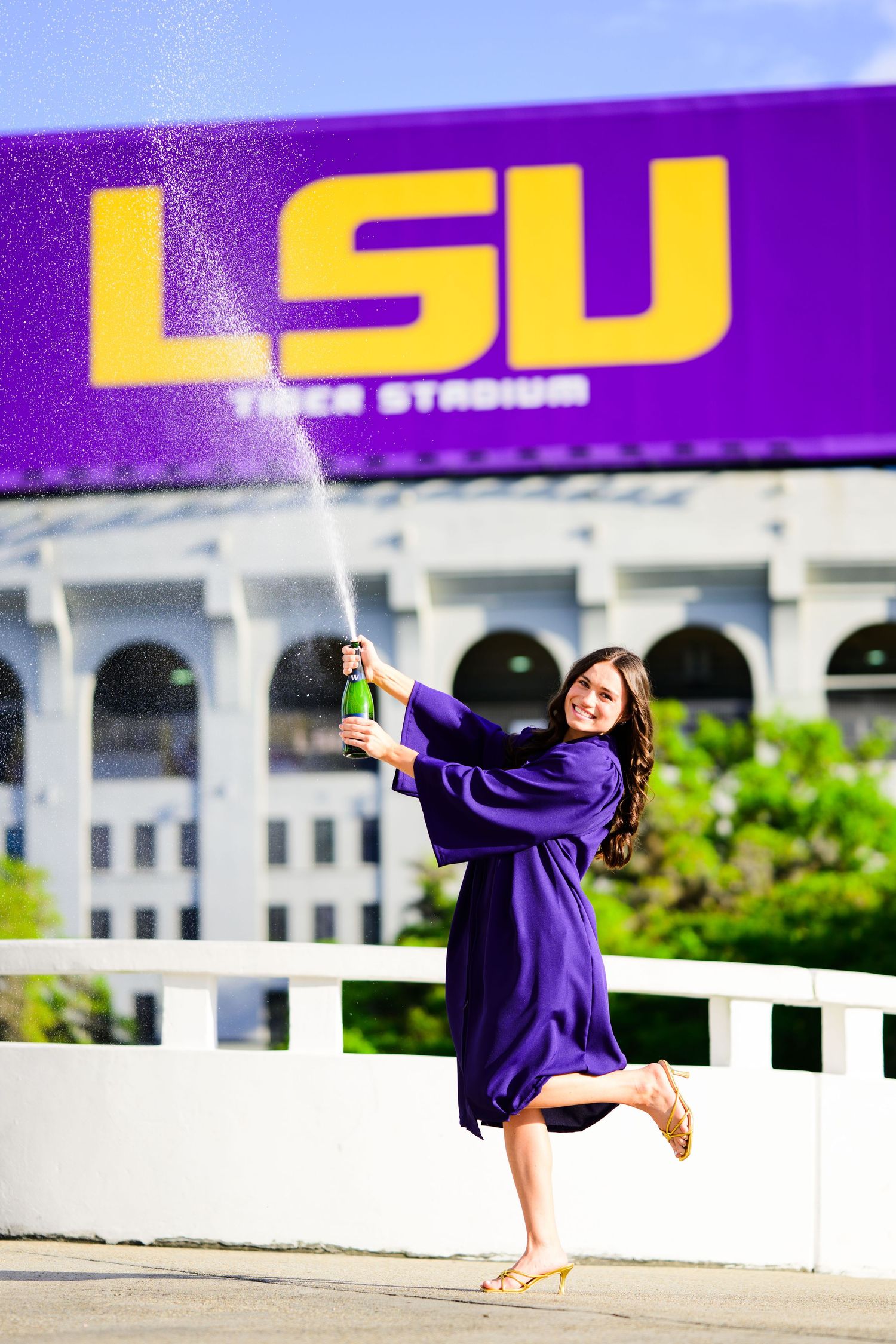 LSU graduate in purple gown sprays champagne bottle in front of Tiger Stadium sign at Louisiana State University.