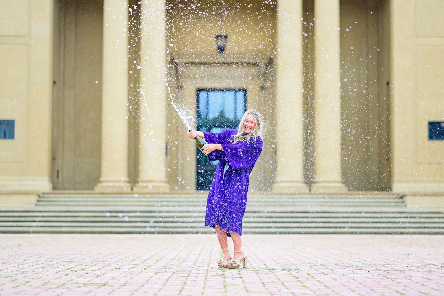 Graduate in purple dress throwing confetti in front of classical building columns.