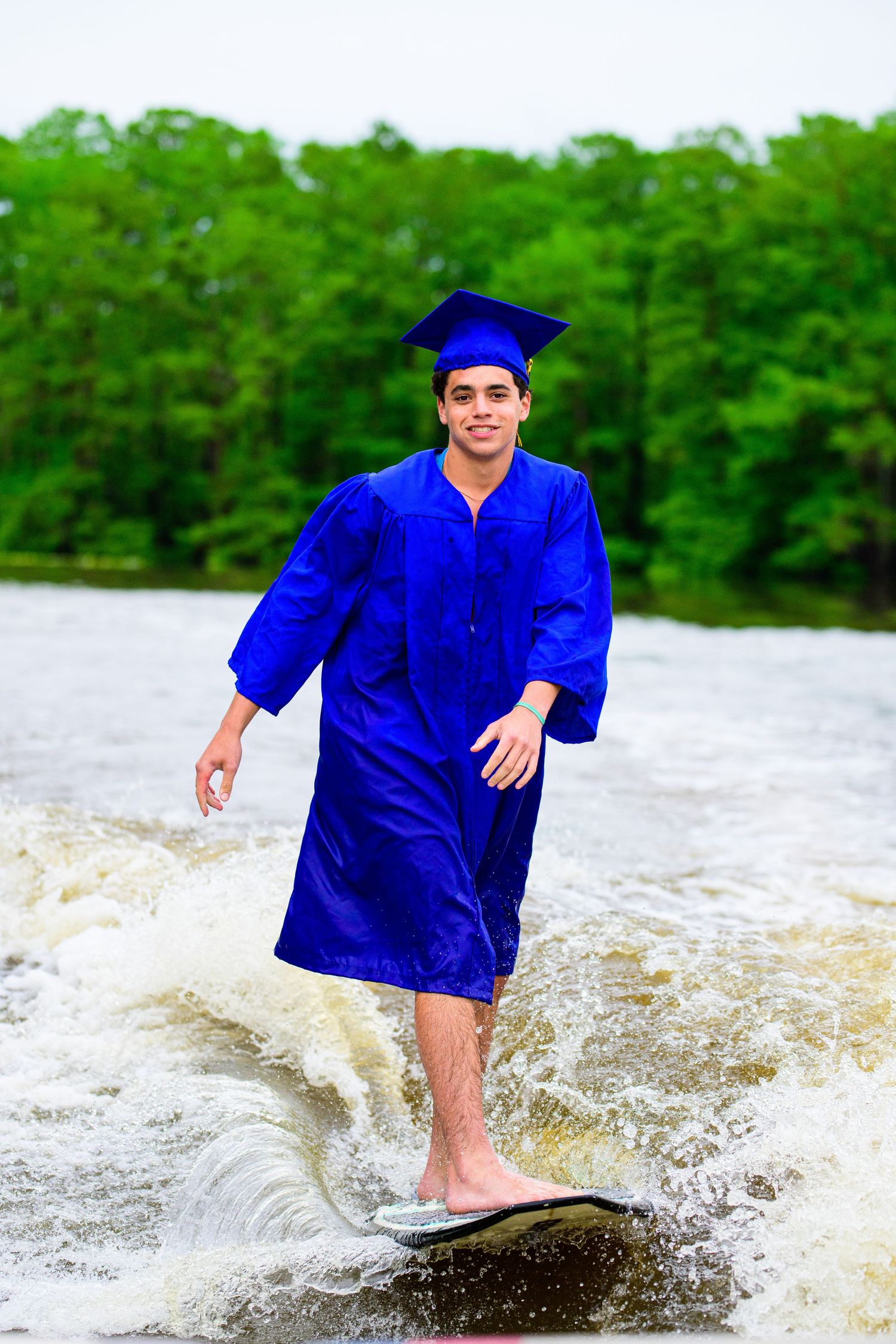 Graduate wakesurfs in full cap and gown on churning lake waters.