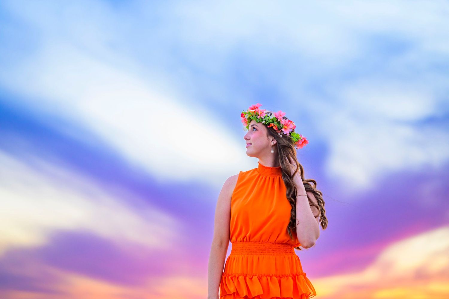 Woman in orange dress with flower crown poses against vibrant sunset sky.
