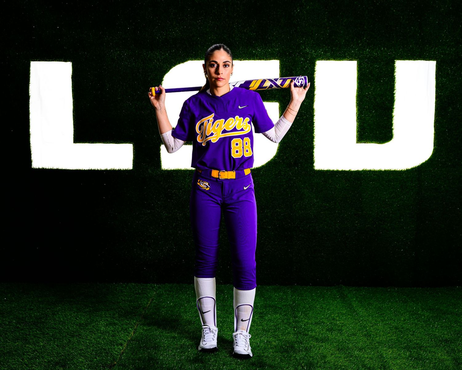 Baseball player wearing purple LSU uniform stands with bat on shoulder in front of large white LSU letters on dark background.