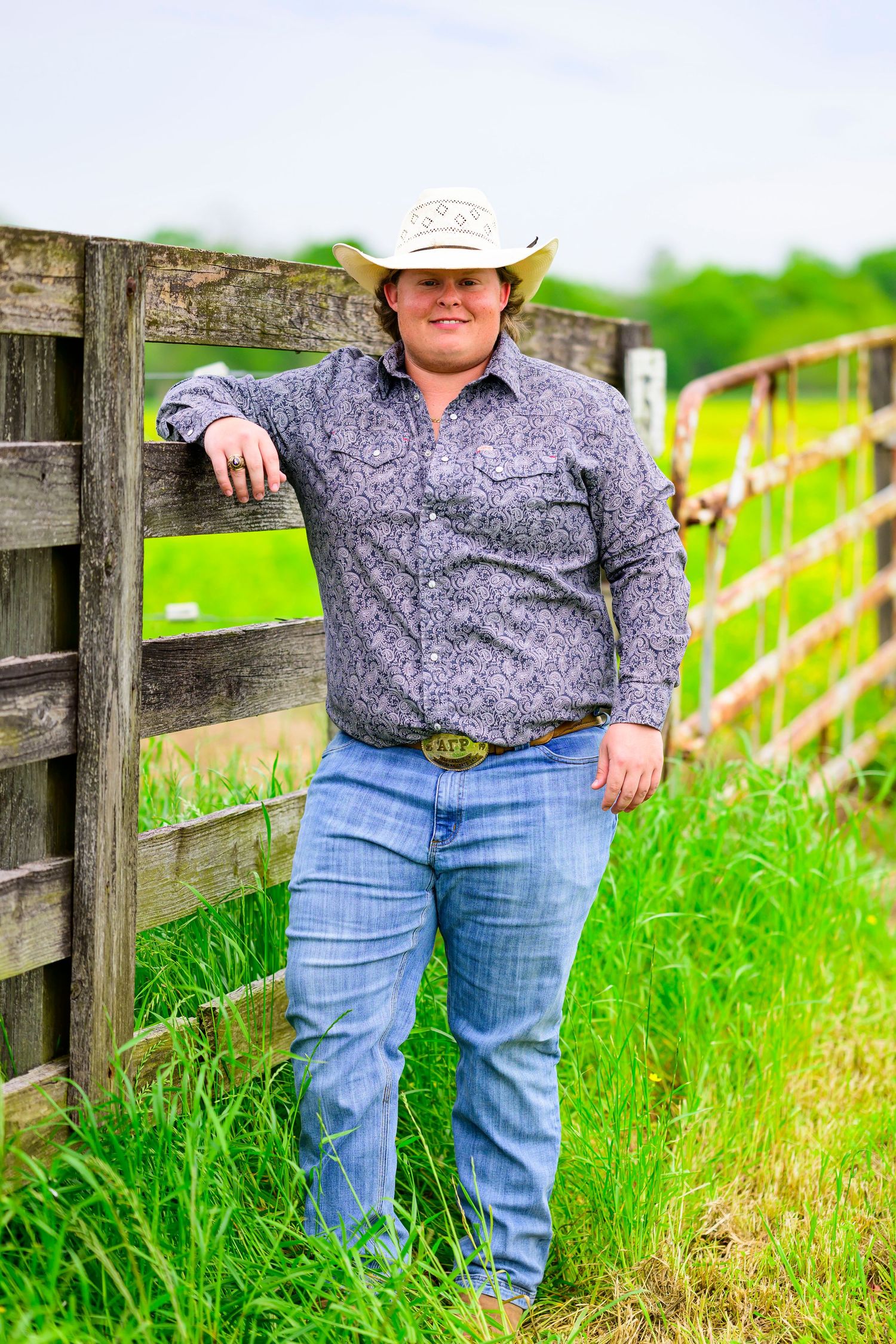 Person in western attire leans against rustic wooden fence on grassy farmland.